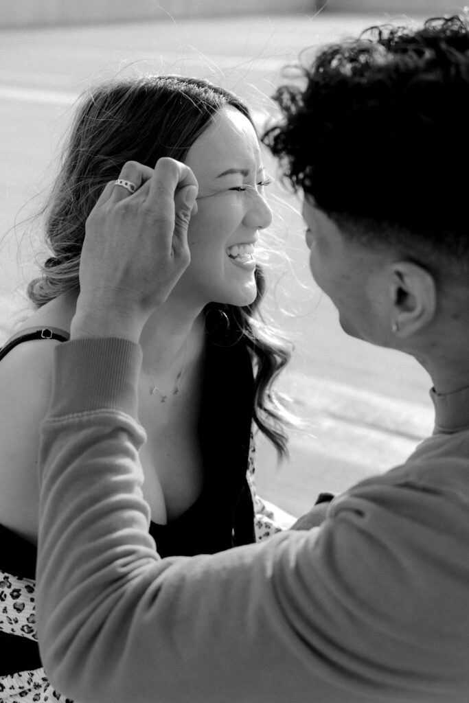 black and white photo of engaged couple laughing together while groom brushes bride's hair back away from her face