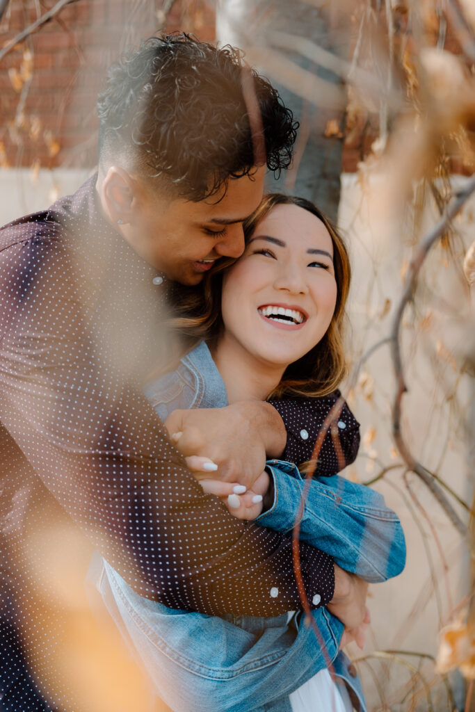 engaged couple under a tree with man hugging woman from behind and laughing together