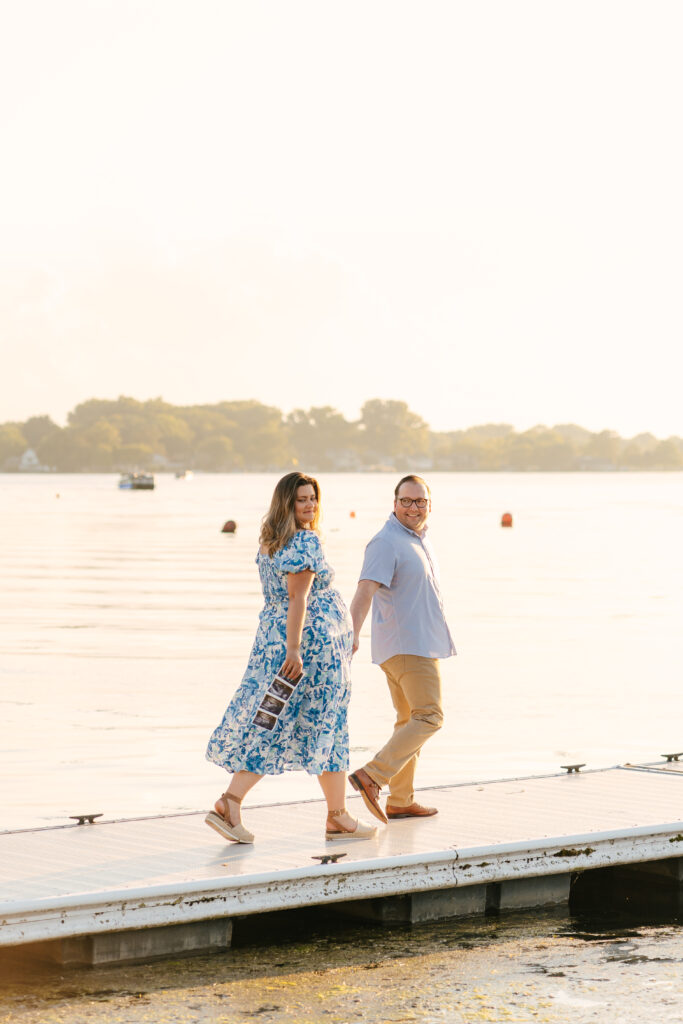 Family photographer capturing natural movement as expecting parents walk along the dock at Winona Lake.